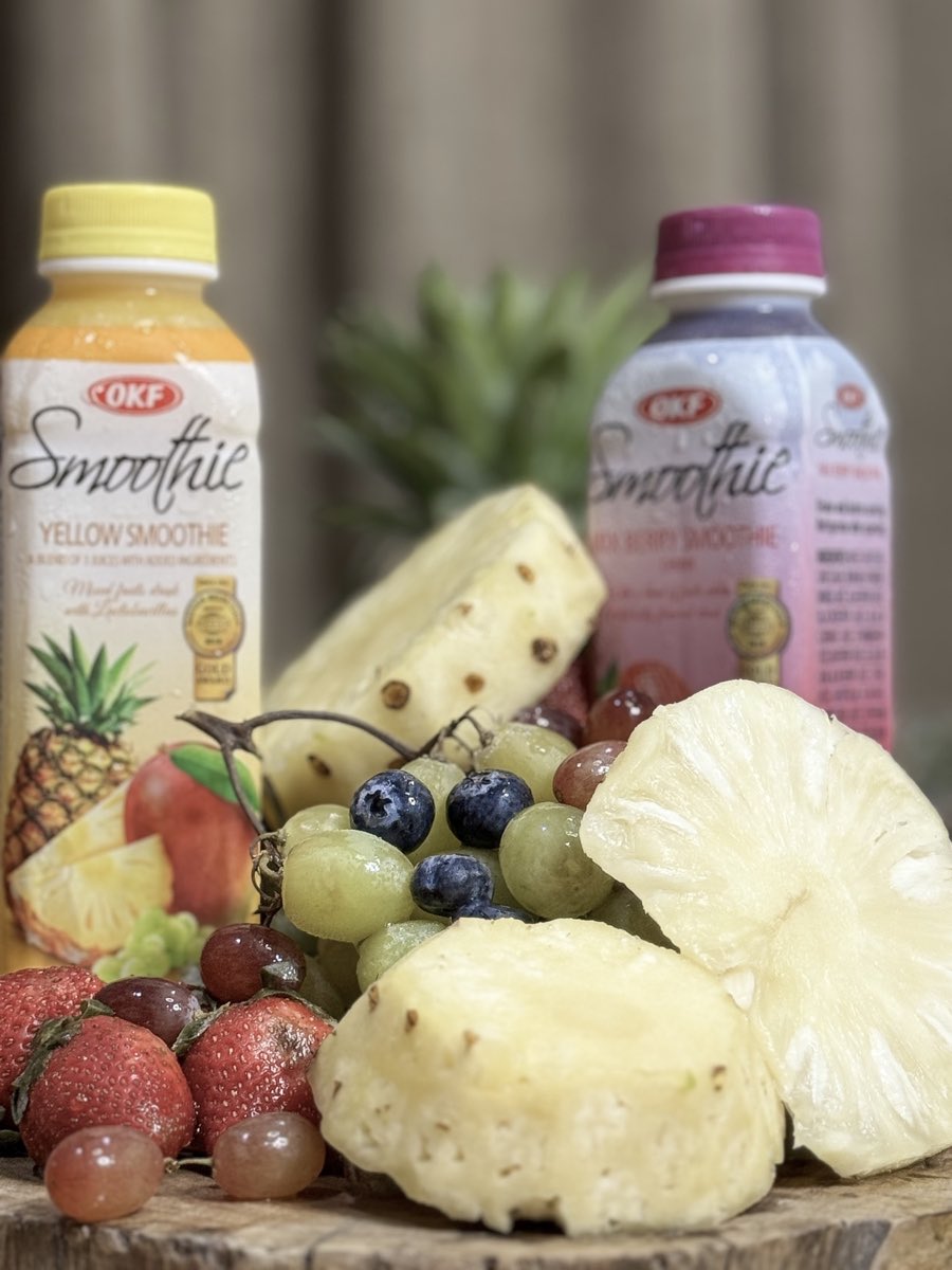 Yellow and mixed berry OKF Korean smoothie bottles surrounded by fresh pineapple, strawberries, grapes and blueberries